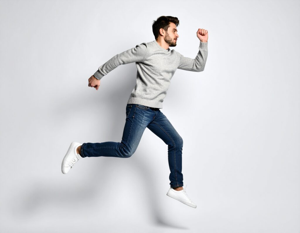 Homme barbu en pull gris et jean sautant avec détermination sur fond blanc.